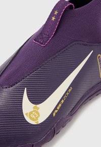 Scarpa da ginnastica viola con una parte superiore in gomma liscia, logo Nike bianco e dettagli dorati. Include una suola testurizzata e cuciture rosa.