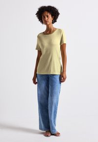 Ljusgul kortärmad topp i mjukt material, ihop med lösa ljusblå denimjeans. Modellen står barfota mot en enkel bakgrund.