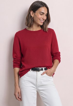 Femme aux cheveux châtain mi-longs, portant une chemise rouge à manches longues rentrée dans un pantalon blanc avec une ceinture noire, souriante et regardant vers le bas.
