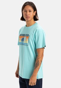 Camiseta de algodón azul claro con un diseño gráfico que incluye palmeras, un sol poniéndose sobre las olas y el logo "Quiksilver" en el centro.