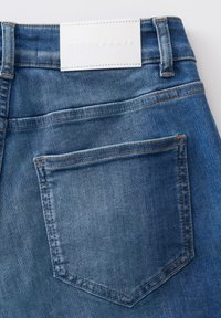 Jeans in denim blu chiaro con tasca posteriore e etichetta in pelle bianca che riporta la dicitura "OPUS PANTS." Cuciture visibili e finitura in tessuto testurizzato.