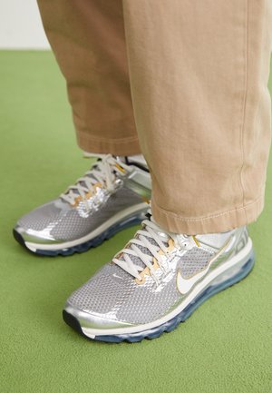 Personne portant un pantalon beige et des baskets argentées Nike Air Max debout sur un tapis vert.