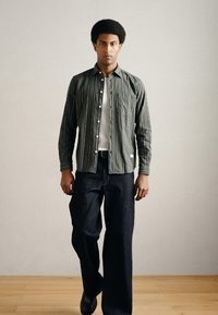 Zielona koszula w paski zapinana na guziki założona na białą koszulkę, zestawiona z ciemnoniebieskimi, szerokimi dżinsami i czarnymi butami. Model stoi na neutralnym tle.