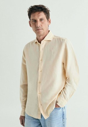 Hombre con cabello corto castaño que lleva una camisa beige abotonada y vaqueros azul claro, de pie con una mano en el bolsillo frente a una pared lisa.