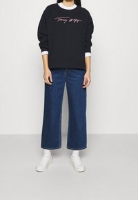 Felpa nera con logo rosa in script, colletto bianco visibile, abbinata a jeans blu a gamba larga e sneakers bianche.