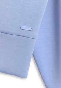 Manche en tissu bleu clair avec une petite plaque métallique rectangulaire embossée "4F" près du poignet.