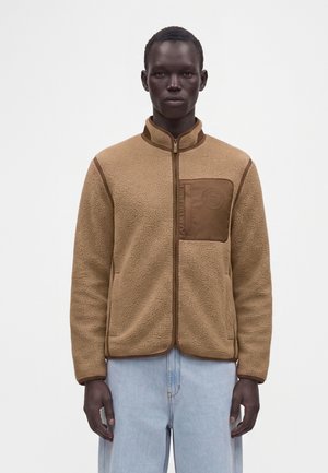 Hombre con chaqueta polar marrón clara con cremallera y bolsillo en el pecho, y vaqueros azul claro, de pie frente a un fondo blanco liso.