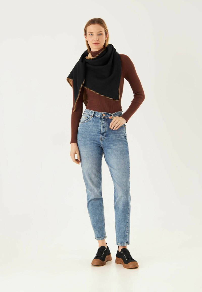 Zwarte sjaal met tan stiksel hangt over een strakke bruine longsleeve, gecombineerd met lichtblauwe hoge taille jeans en zwarte sneakers.