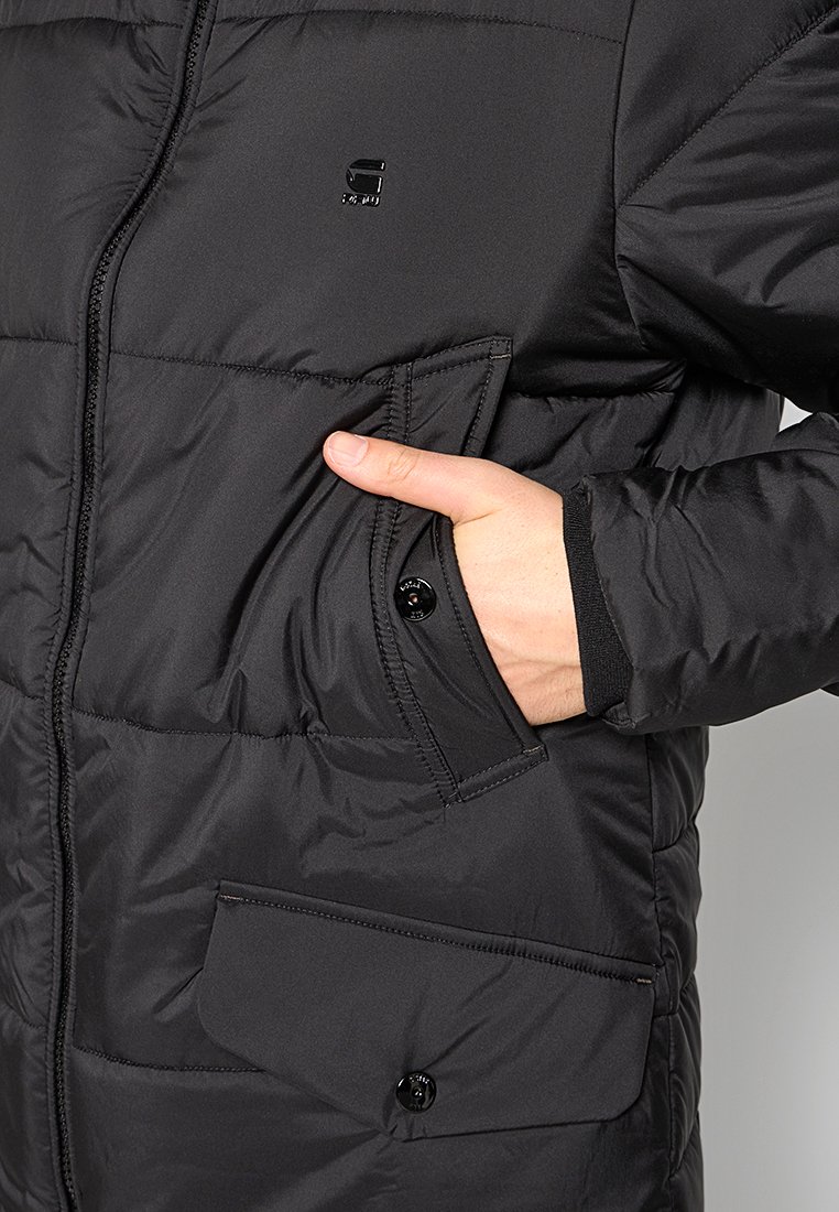 Chaqueta negra aislante con cierre de cremallera, un logotipo brillante en el pecho y dos bolsillos con botones a presión. Textura suave y acolchada.