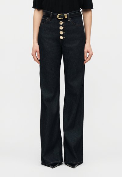 Γυναίκα που φοράει ψηλόμεσο σκούρο μπλε τζιν με φαρδιά πόδια, πέντε χρυσά κουμπιά και μαύρη ζώνη με χρυσή αγκράφα.