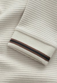 Tissu en maille gaufrée couleur crème avec un poignet arborant une rayure horizontale marron et noire. Surface texturée avec une finition douce.