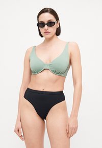 Top bikini verde a coste con spalline larghe e dettaglio centrale a nodo, abbinato a slip bikini neri a vita alta. Texture liscia, design minimalista.