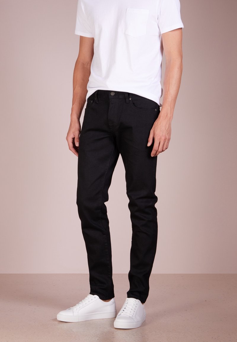 Black denim jeans with a slim fit, five pockets, and a button-fly closure, paired with a white T-shirt and white sneakers.