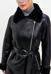 Černý kabát s lesklým povrchem, velkým kožíškovým límcem a stříbrnými zipy. Součástí je pásek v pase a postranní kapsy na zip.