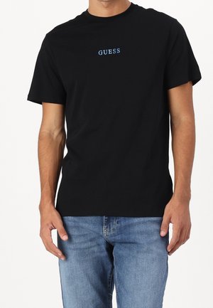 Homme portant un t-shirt noir à manches courtes avec un texte bleu "GUESS", associé à un jean bleu en denim sur un fond uni.