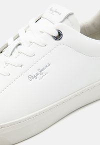 Baskets basses blanches avec lacets blancs, logo Pepe Jeans London argenté sur le côté et œillet métallique rond près du trou supérieur du lacet.