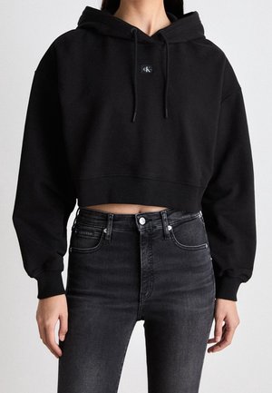 Kobieta w czarnej, krótkiej bluzie z kapturem i troczkami oraz w wysokim stanem ciemnoszarych dżinsach, stojąca na jednolitym tle.