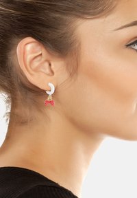 Orecchino ad anello bianco con un piccolo distintivo a fiocco rosso, caratterizzato da una texture lucida. Il design pende dal lobo, mostrando uno stile moderno.