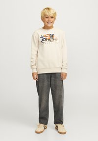 Dečko nosi krem majicu s logom "JACK & JONES" u narančastoj i plavoj boji, u kombinaciji s tamnosivim trapericama i bež tenisicama.
