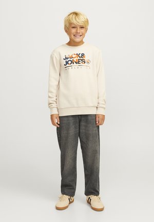 Poika, jolla on yllään kermanvärinen huppari, jossa on oranssi ja sininen "JACK & JONES" -logo, yhdistettynä tummanharmaisiin farkkuihin ja beigeihin lenkkareihin.