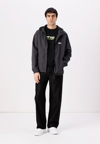 Dunkelgraue, leichte Jacke mit Kapuze, Frontreißverschluss und seitlichen Taschen, über einem schwarzen T-Shirt mit grünem Text. Schwarze Hose und weiße Sneaker.