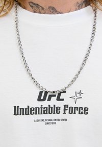 Person med en sølv kædereg necklace og en hvid t-shirt med teksten "UFC Undeniable Force Las Vegas, Nevada, United States Siden 1993".