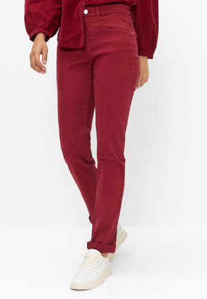 Person iført røde slim-fit bukser og matchende rød langærmet skjorte, parret med hvide sneakers, stående mod en hvid baggrund.