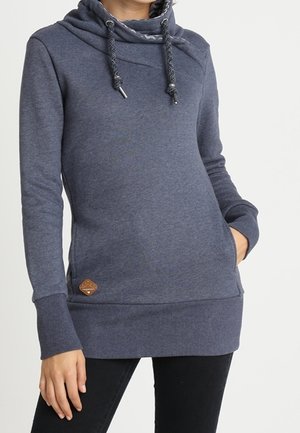 Femme portant un sweat à capuche bleu marine à manches longues avec cordons et une poche avant, associé à un pantalon noir.