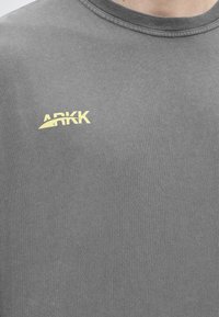 Κοντινή λήψη γκρι πουκαμίσου με κίτρινο λογότυπο ARKK τυπωμένο στο πάνω αριστερό μέρος του στήθους.