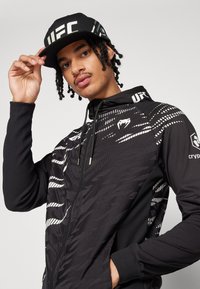 Črna zip-up jakna z abstraktnimi belimi vzorci, dolgimi rokavi in kapuco, v kombinaciji s črnim klobukom z logotipom UFC.