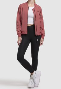 Femme portant une veste bomber rose poudré, un haut court blanc, des leggings noirs et des baskets montantes blanches, debout devant un fond uni.