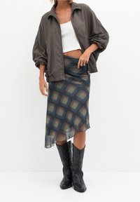 Vrouw in een grijze oversized jas, witte cropped top, asymmetrische geruite rok en zwarte cowboylaarzen, staand tegen een witte achtergrond.