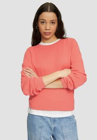 Sweatshirt mit Korallenstruktur, langen Ärmeln, geripptem Kragen und Bündchen, über einem weißen Hemd getragen. Kombiniert mit hellblauen Jeansshorts.