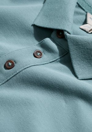 Atvaizdas iš arti: žalsvai mėlynos faktūruotos polo marškinėlių audinys su plokščiu apykakliu ir trimis rudos spalvos mygtukais ant sagos juostos.