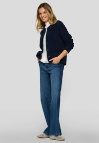 Marineblauer, zum Knöpfen gearbeiteter Cardigan aus weichem Stoff, getragen über einem weißen Oberteil, kombiniert mit ausgestellten blauen Jeans und metallischen Schuhen.