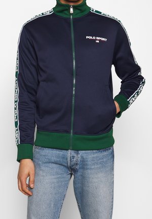 Osoba ubrana w granatowo-zieloną kurtkę Polo Sport zapinaną na zamek z logo na piersi i rękawach, w połączeniu z jasnoniebieskimi jeansami.