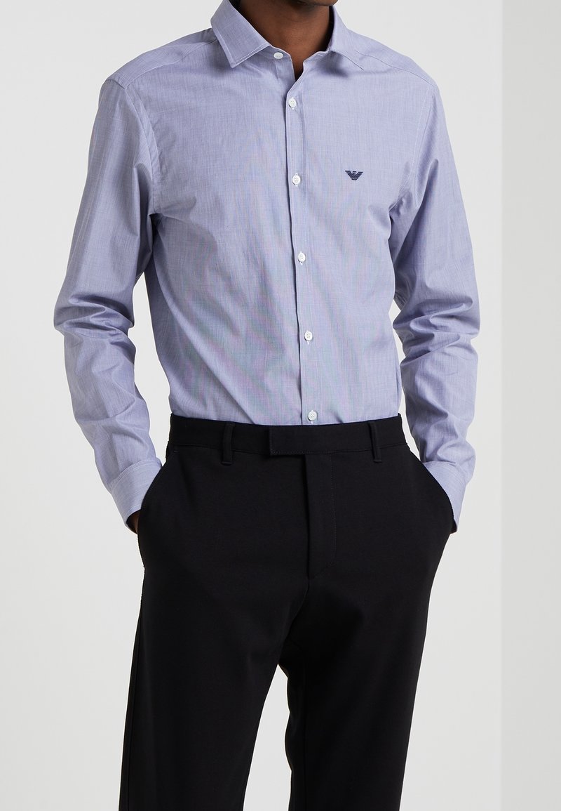 Chemise boutonnée bleu clair avec un petit logo foncé sur la poitrine, associée à un pantalon noir ajusté. Matière en coton, texture lisse.