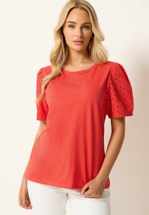 Mujer rubia vistiendo una blusa coral con mangas abullonadas con ojales y pantalones blancos, de pie frente a un fondo claro y liso.