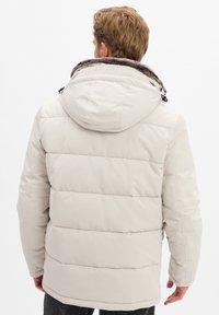 Man in een beige gewatteerde jas met een met bont gevoerde capuchon, gezien van achteren tegen een effen witte achtergrond.