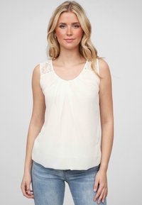 Blouse blanche sans manches avec des détails en dentelle aux épaules, devant plissé, tissu doux, encolure arrondie et coupe décontractée, associée à un jean bleu clair.