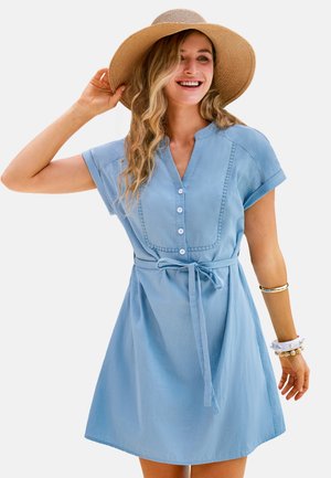 Mujer sonriente con un vestido azul de manga corta con botones y cinturón, complementado con un sombrero de paja y pulseras en su muñeca derecha.