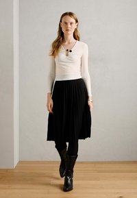 Haut blanc à manches longues avec un détail en clé de serrure, associé à une jupe plissée noire et des bottes noires jusqu'aux genoux, sur un fond neutre.