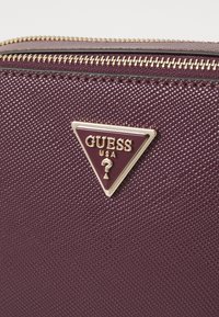 Burgunderfarbenes, strukturiertes Materialportemonnaie mit goldenen Beschlägen. Verfügt über eine dreieckige Logoschild mit "GUESS" und einem Fragezeichen-Design. Mit Reißverschluss.