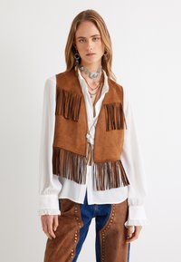 Gilet in suede marrone con frange e finitura liscia, indossato sopra una camicia bianca. Presenta nappine lungo l'orlo e perline decorative sui pantaloni.