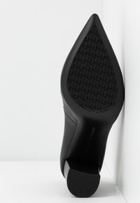 Chaussure à talon haut noire avec un bout pointu, une tige en cuir lisse, une semelle extérieure en caoutchouc noir texturé et un logo embossé sur le talon.