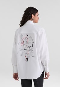 Camicia bianca con bottone, dal taglio morbido, con un design di schizzo disegnato a mano sul retro in bianco e rosso, e maniche lunghe.
