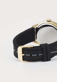 Bracelet de montre en silicone noir avec une surface texturée, dotée d'une boucle en métal doré et d'une bande grise. L'arrière de la montre est argenté avec une gravure.