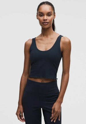 Slank kvinde med mørk hud og samlet hår iført en sort cropped tanktop og sorte leggings med høj talje, stående mod en ensfarvet lys baggrund.