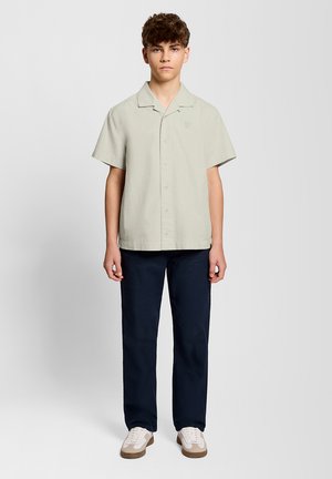 Teenager med krøllet hår iført en lysebeige kortærmet skjorte med knapper, mørkeblå bukser og beige sneakers, stående foran en hvid baggrund.