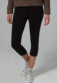 Zwarte capri leggings van rekbaar materiaal, met een strakke pasvorm die net onder de knie eindigt, gecombineerd met witte sneakers.